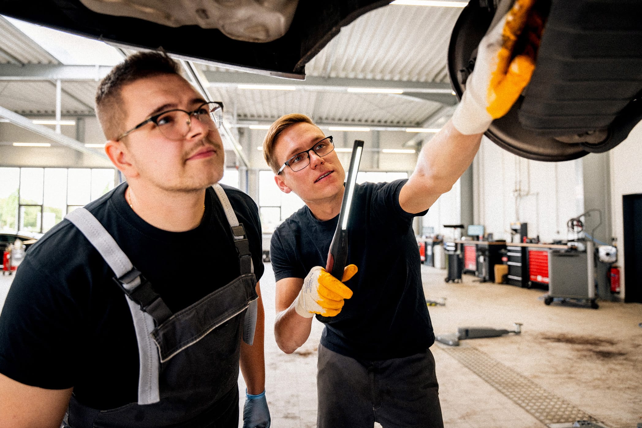 Zwei Männer in Arbeitskleidung, die den Unterboden eines Autos auf einer Hebebühne begutachten. Einer der beiden Männer hält eine Werkstattlampe in der Hand und deutet auf ein Bauteil am Fahrzeug.
